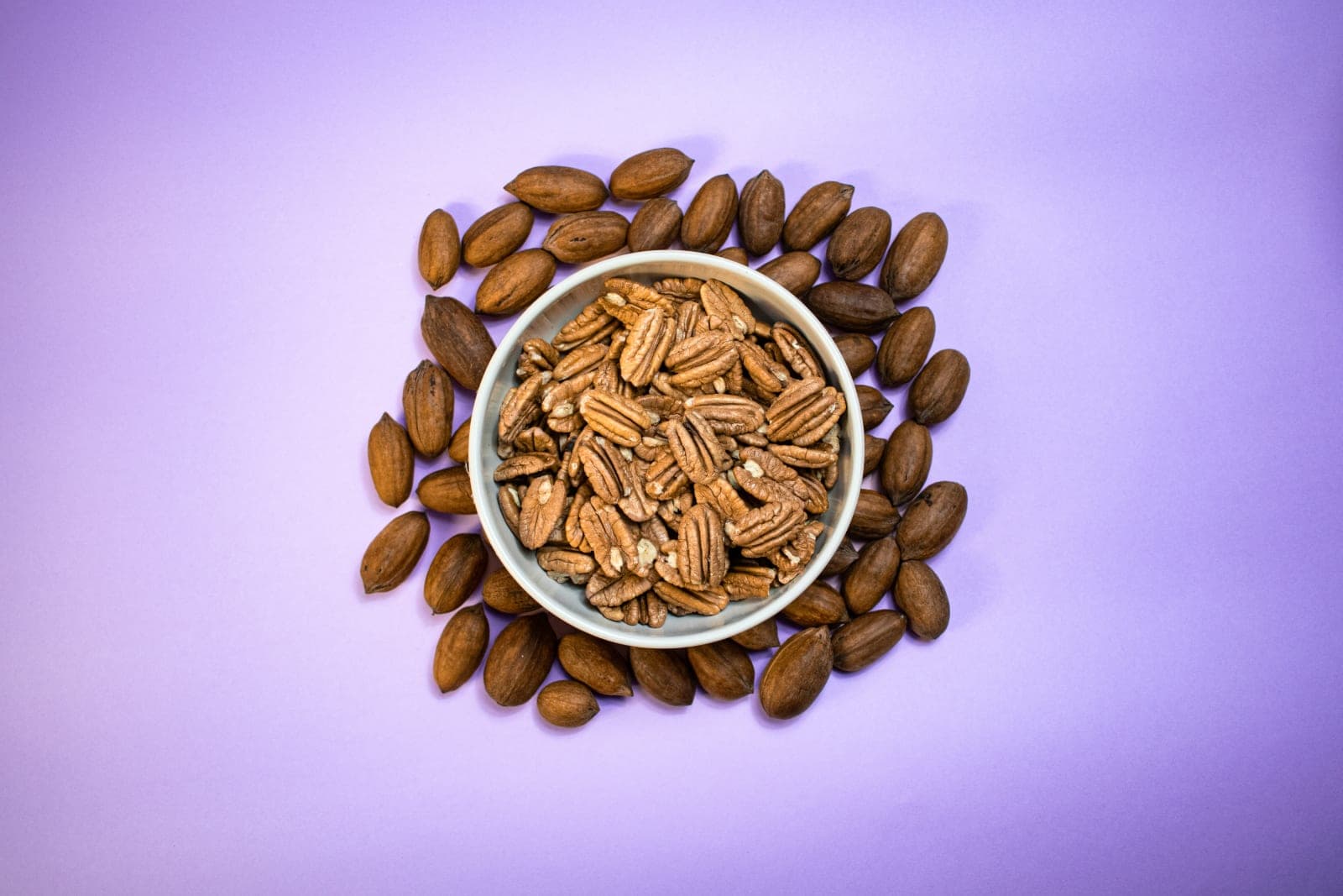 Nueces pecanas peladas en un tazón, rodeadas de nueces con cáscara